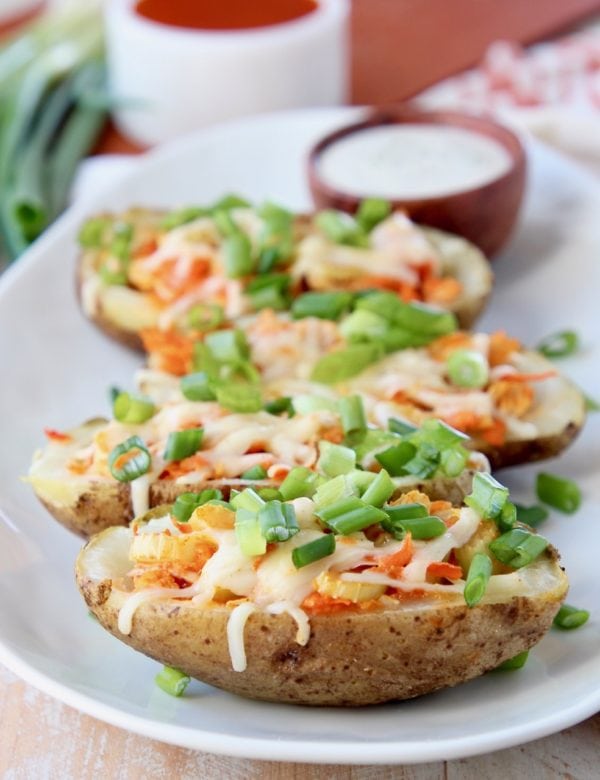 Buffalo chicken potato skins lined up on a white oval plate topped with diced green onions with a small bowl of ranch dressing in the background