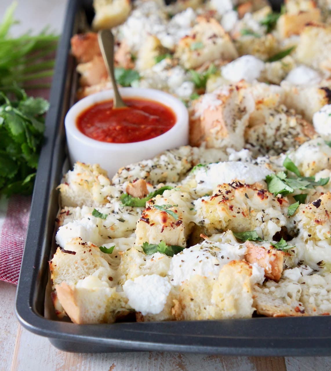 Cubed garlic bread, covered in cheese and fresh parsley on a baking sheet with a small bowl of marinara sauce