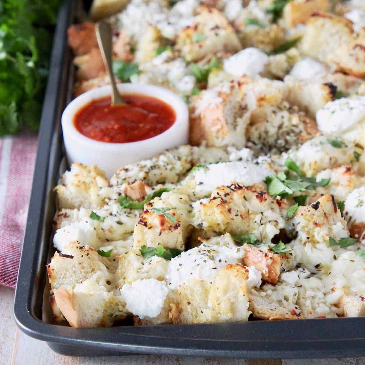 Cubed garlic bread covered in cheese on a baking sheet with a side of marinara sauce in a white bowl with a small spoon