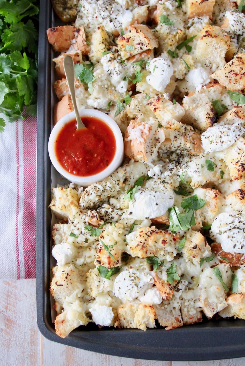 Cheesy garlic bread nachos on a baking sheet with a small bowl of marinara sauce in the corner with a spoon in the bowl