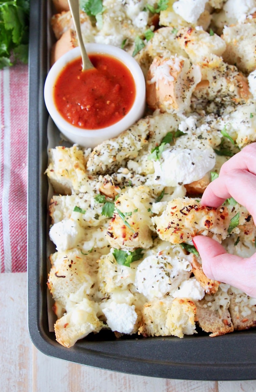 Hand grabbing a piece of garlic bread nachos off of a baking sheet with a small bowl of marinara sauce in the corner