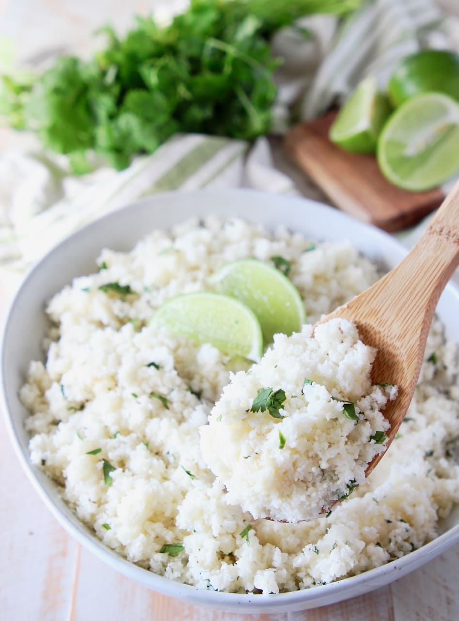 Wooden spoon of cilantro lime cauliflower rice coming out of a bowl of cauliflower rice with lime wedges on top and fresh cilantro in the background