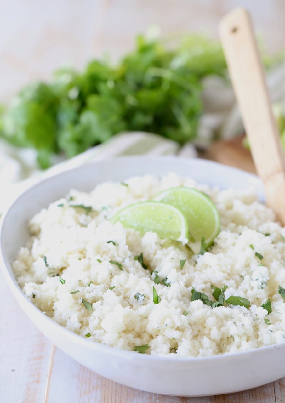 Cilantro lime cauliflower rice in white bowl with lime wedges on top and wooden spoon in bowl, with fresh cilantro in background