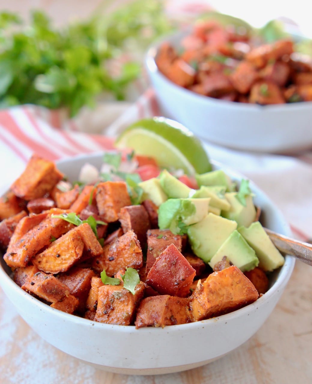 Diced roasted sweet potatoes and diced avocado in quinoa bowl with lime wedge and fork, with orange and white striped towel underneath and cilantro in background