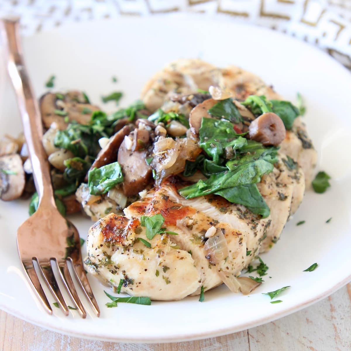 White plate of sliced spinach mushroom chicken with copper fork on the side of the plate, sitting on a gold and white patterned napkin