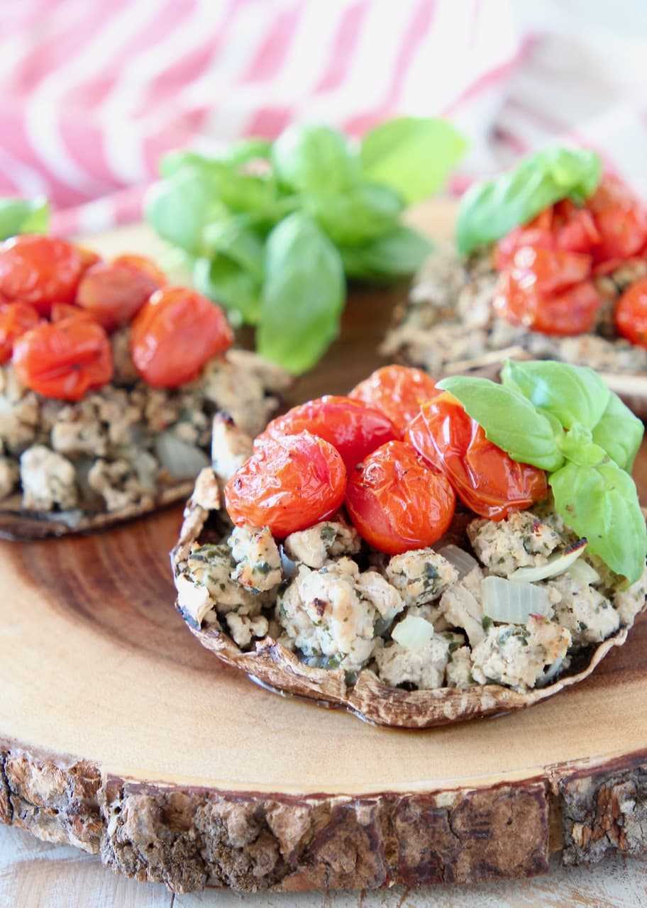 Stuffed portobello mushrooms filled with ground turkey, cherry tomatoes and fresh basil leaves sitting on round wood cutting board with red and white striped towel in background