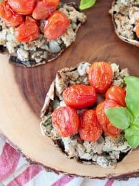 Overhead shot of stuffed portobello mushrooms filled with ground turkey, red cherry tomatoes and fresh basil leaves, sitting on a wood cutting board on top of a red and white striped towel