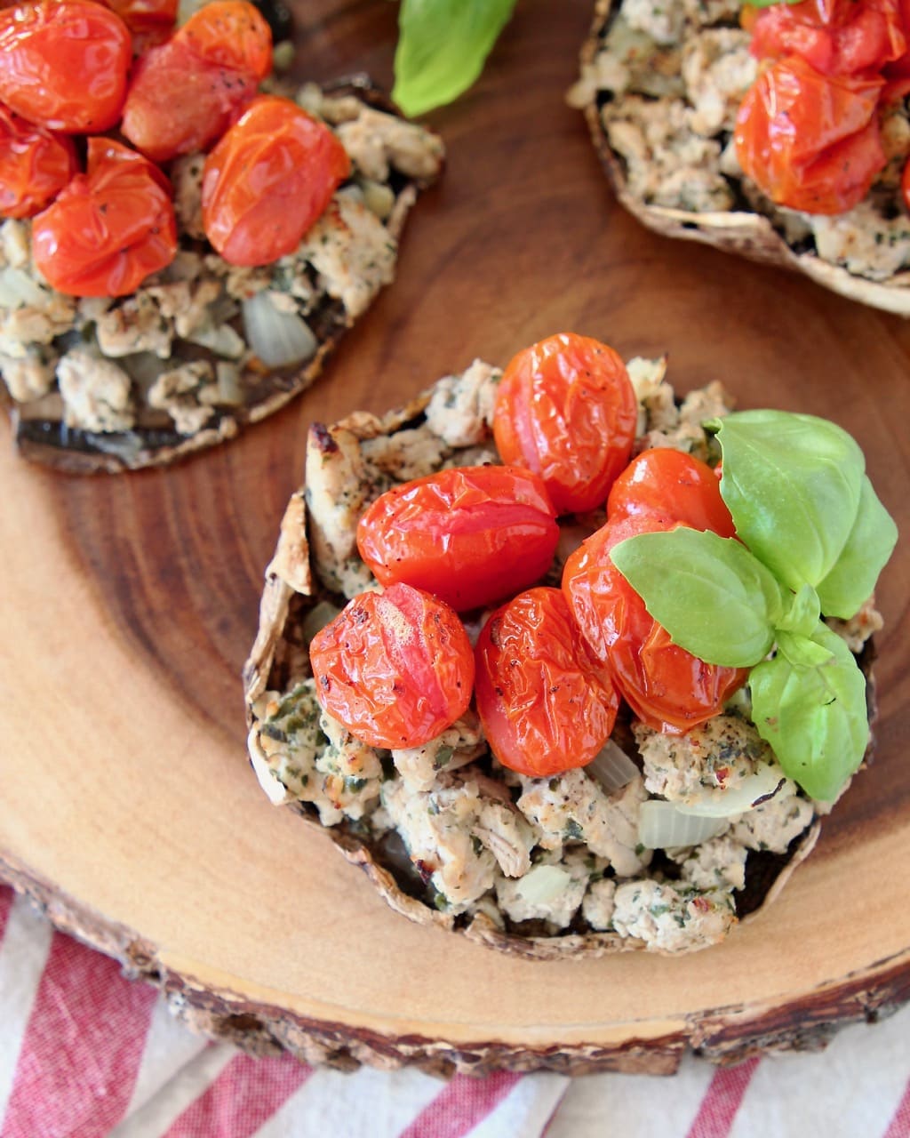 Overhead shot of stuffed portobello mushrooms filled with ground turkey, cherry tomatoes and fresh basil leaves, sitting on a round, raw edge wood cutting board, sitting on a red and white striped towel
