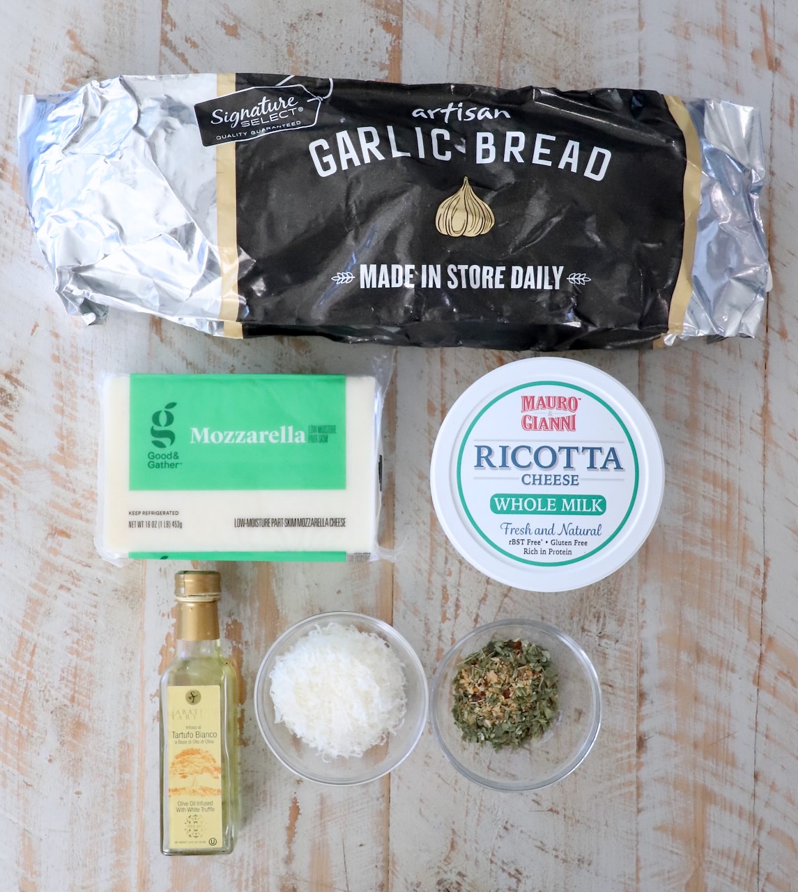Packaged garlic bread on a wooden surface, surrounded by mozzarella, ricotta, olive oil, grated cheese, and herbs