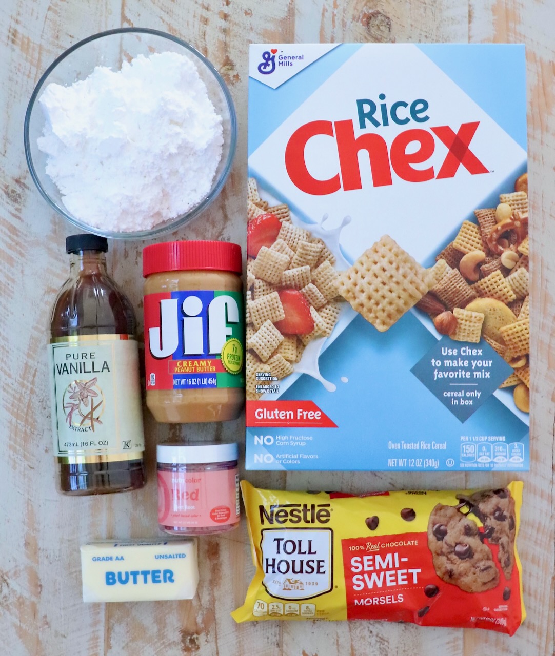 Ingredients for a snack mix are arranged on a wooden surface: a box of Rice Chex, peanut butter, vanilla extract, powdered sugar, butter, chocolate chips, and red powdered food coloring