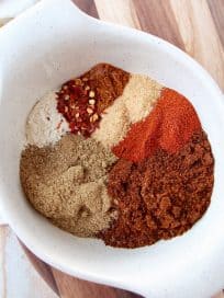 Eight spices for chili seasoning in a white bowl on a wood cutting board