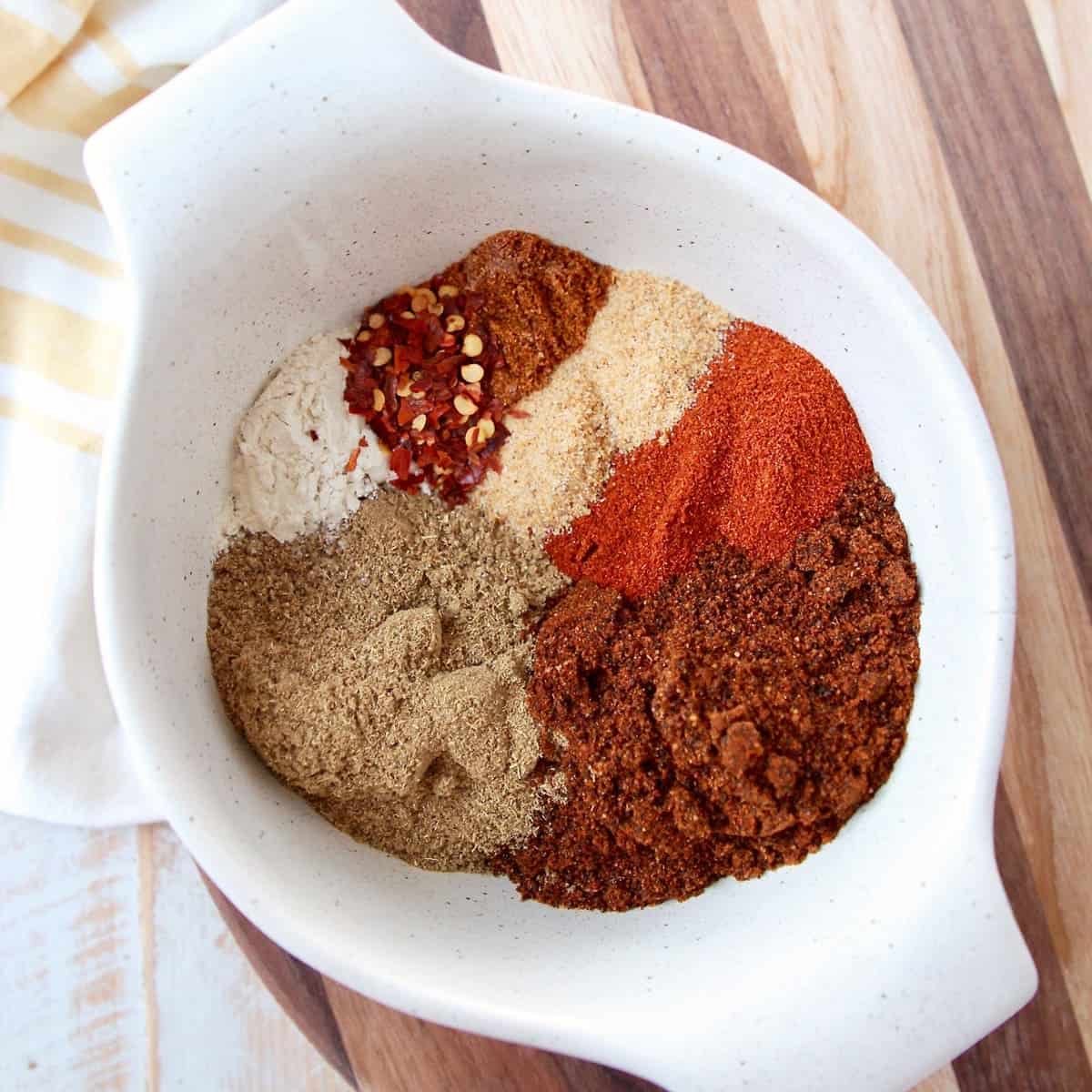 Eight spices for chili seasoning in a white bowl on a wood cutting board