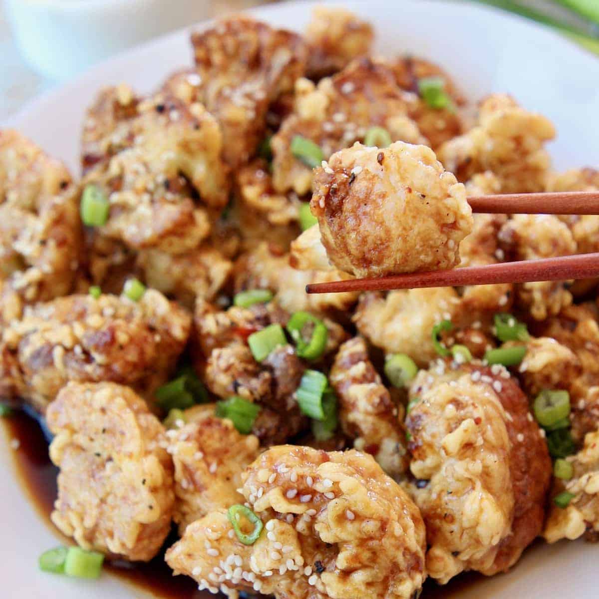 Piece of fried cauliflower in chopsticks, held over plate of fried cauliflower with Korean BBQ sauce