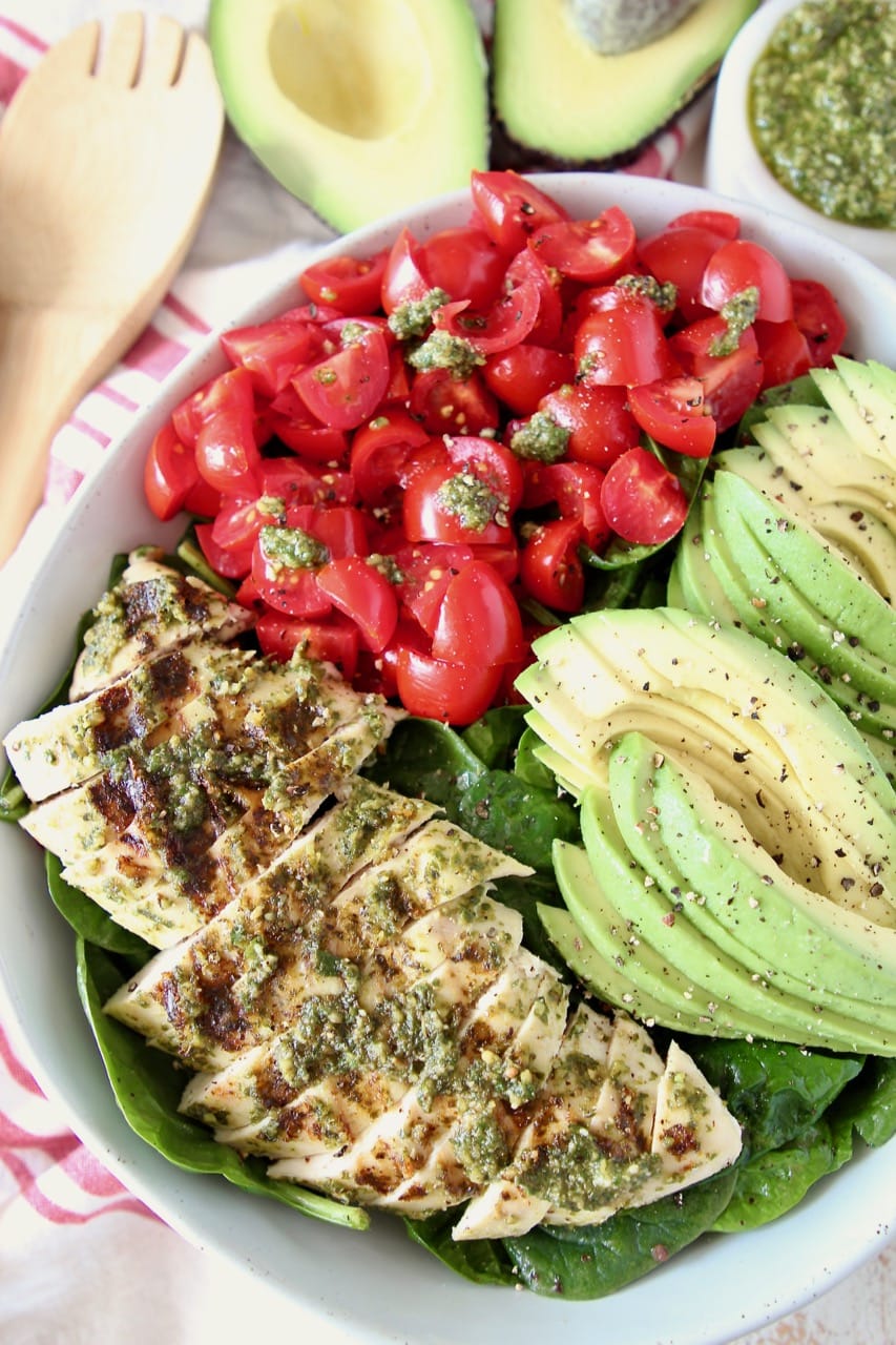 Sliced pesto chicken, sliced avocado and diced cherry tomatoes on top of spinach salad in bowl