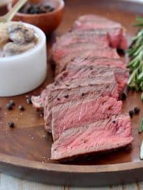 Sliced wagyu strip steak on wood tray with rosemary sprigs and black peppercorns