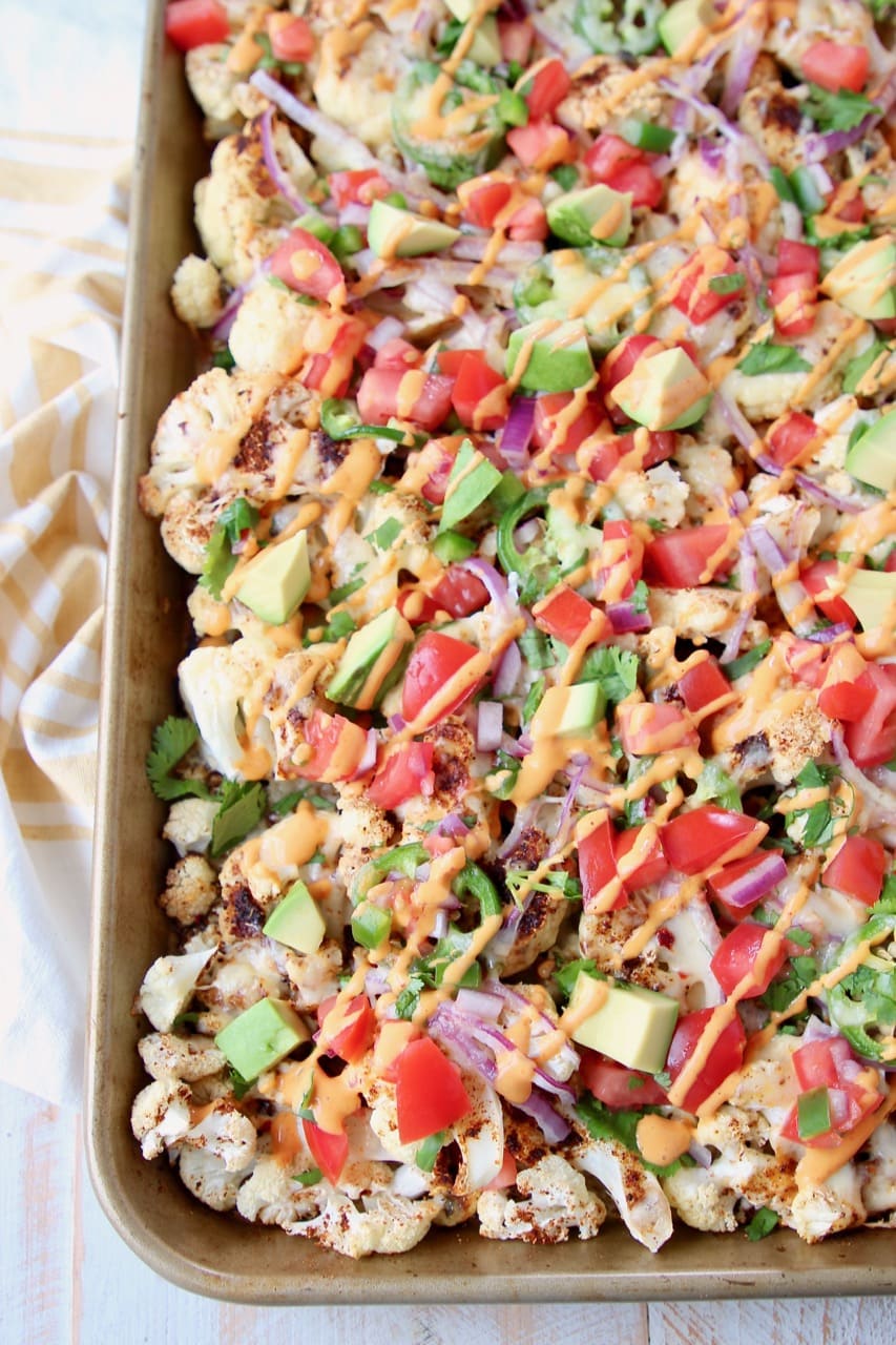 Overhead shot of cauliflower nachos on a baking sheet with chipotle cream sauce drizzled on top