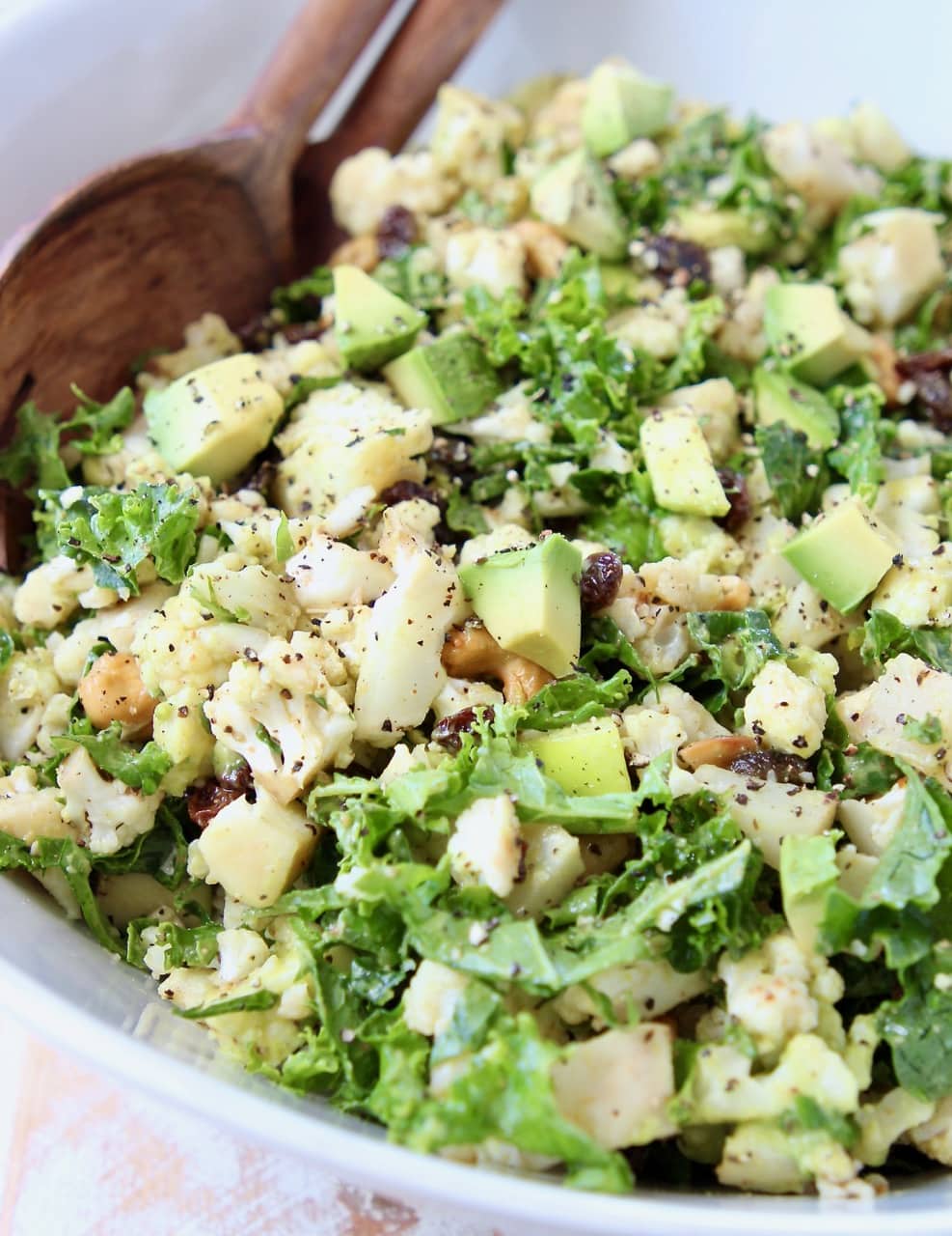 Cauliflower salad with kale, raisins and avocado in white bowl with brown wooden spoon