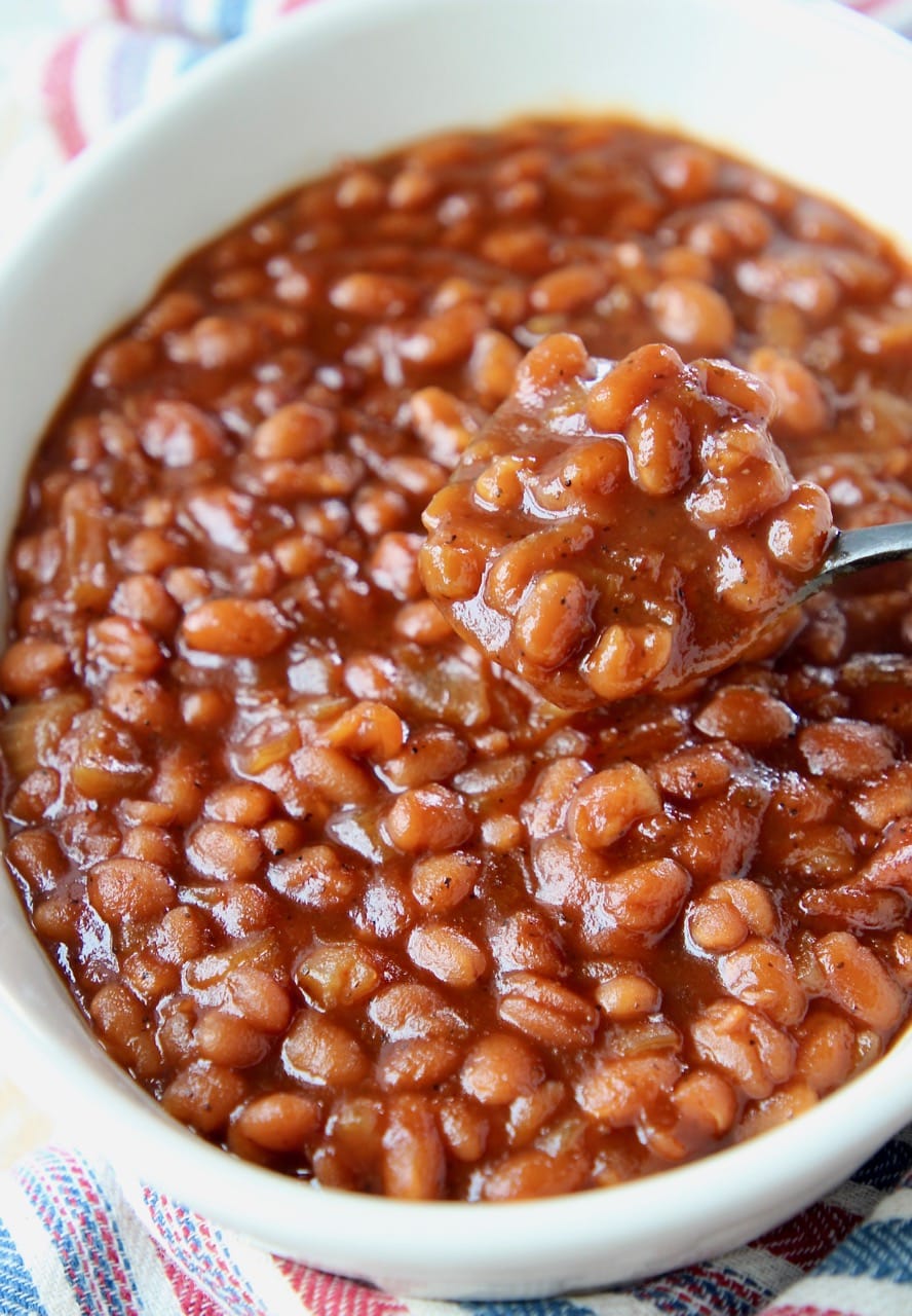 Baked beans in white bowl with black and silver spoon lifting beans out of the bowl