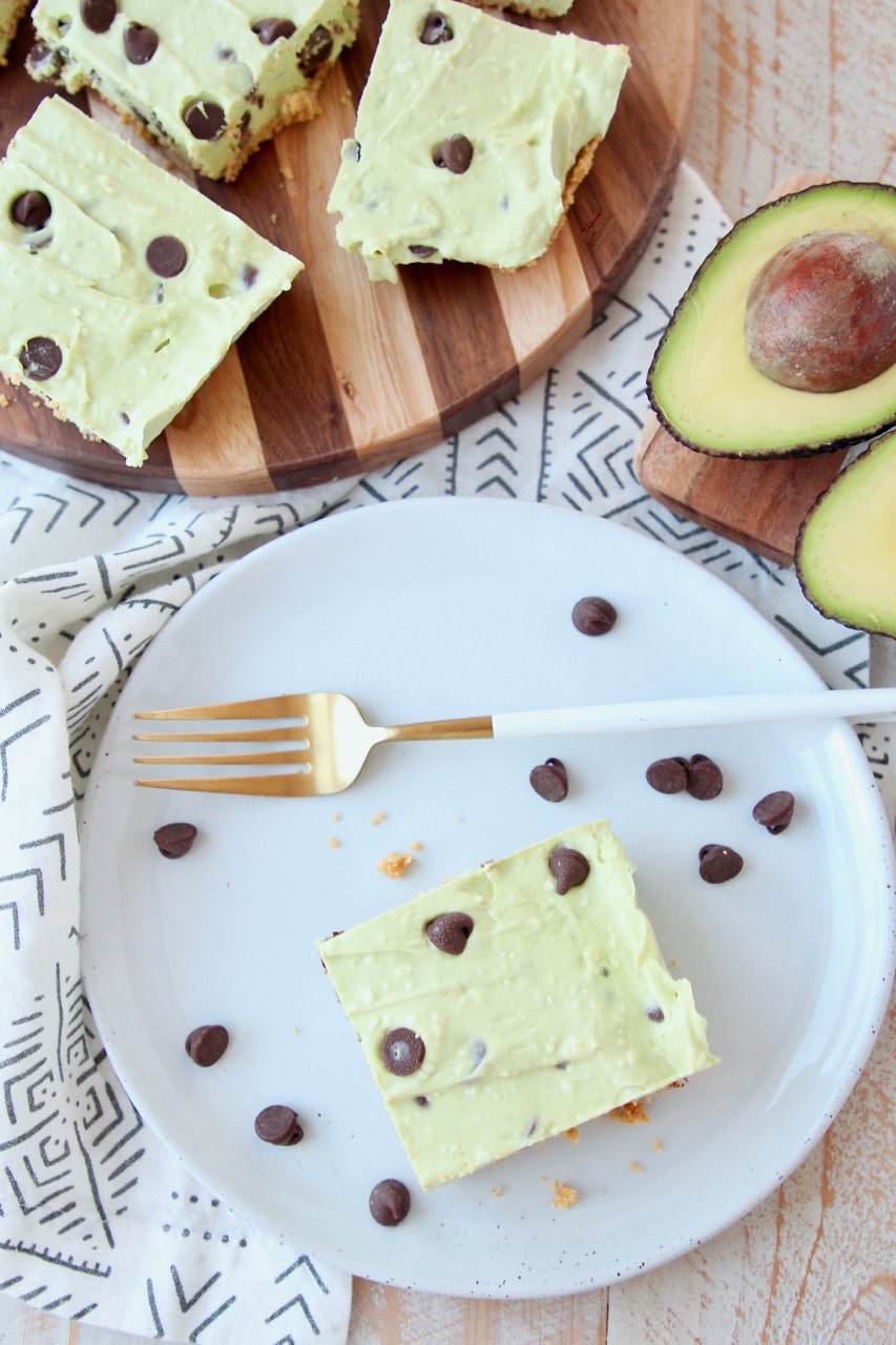 Overhead shot of avocado cheesecake bars on plate and cutting board with chocolate chips and halved avocado