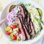 Grilled steak on top of salad in a white bowl with gold salad serving spoons