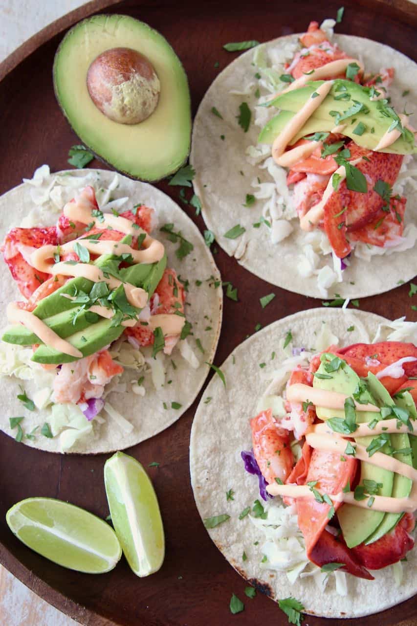 Overhead image of lobster tacos on wood tray with lime wedges and half an avocado