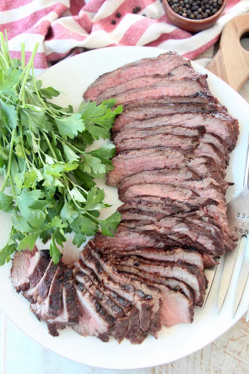 Sliced tri tip steak on white plate with fresh parsley