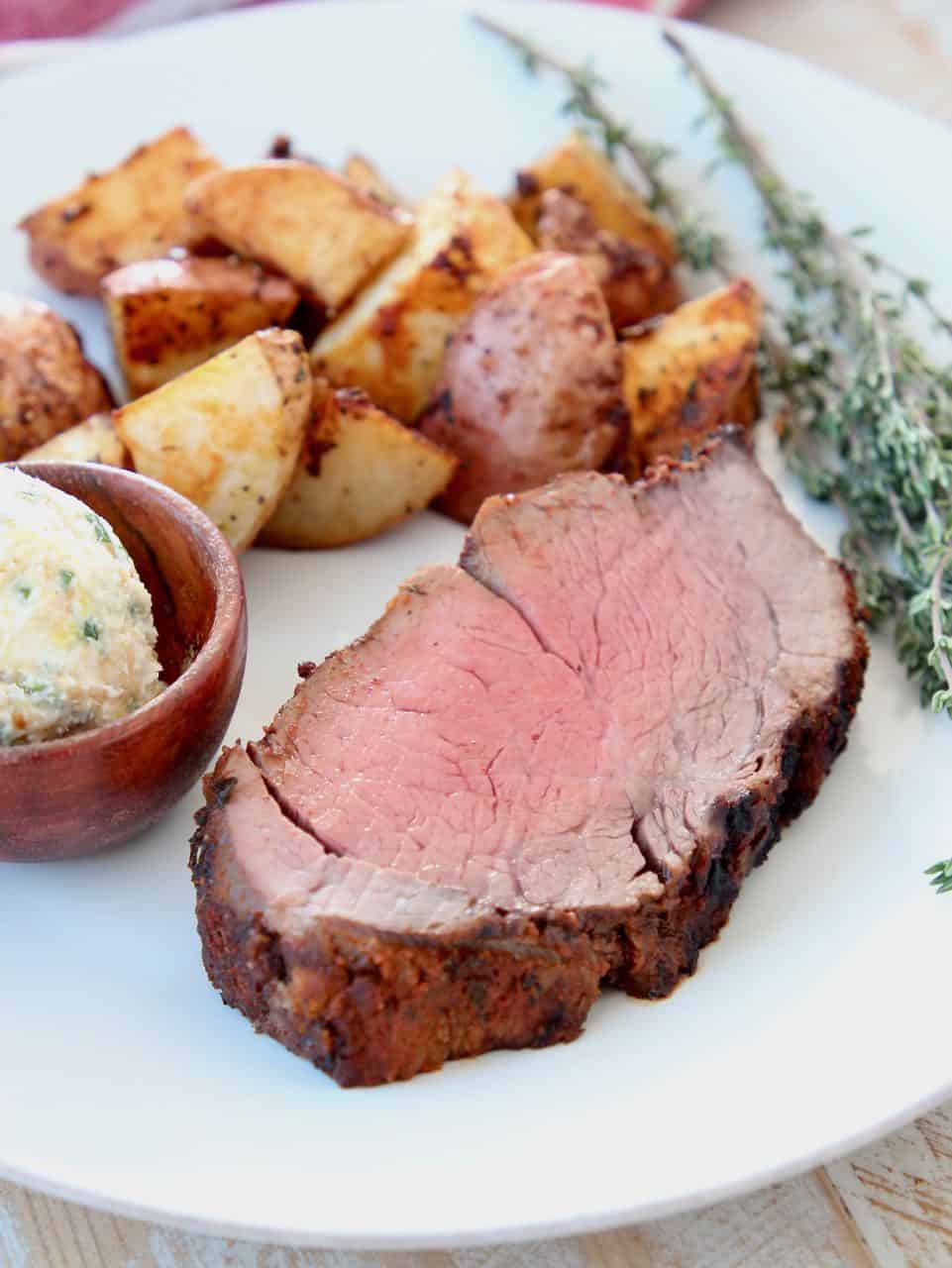Thick sliced beef tenderloin on plate with herbs, potatoes and butter