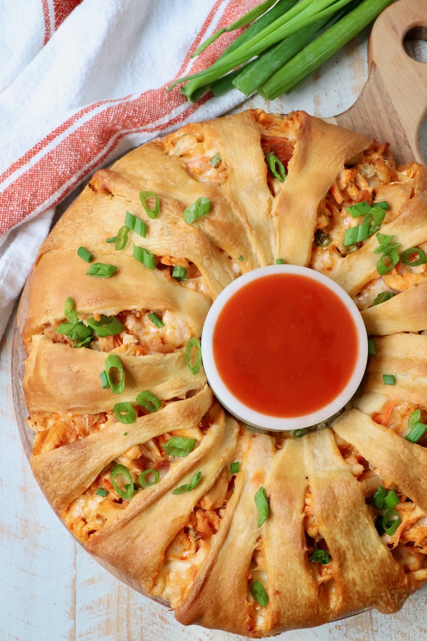 buffalo chicken crescent roll ring on a wood serving tray with a bowl of buffalo sauce in the middle