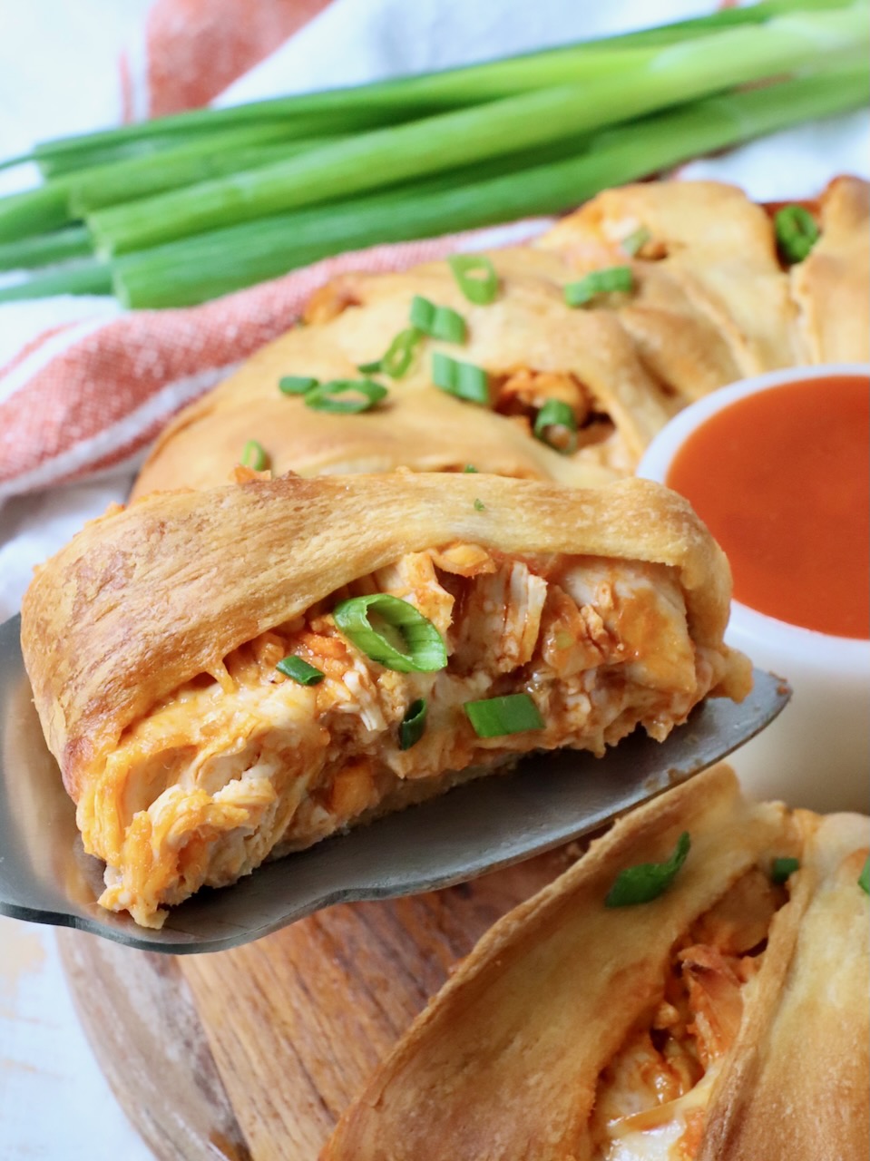 slice of buffalo chicken crescent ring on a spatula with green onions and buffalo sauce in a bowl on the side