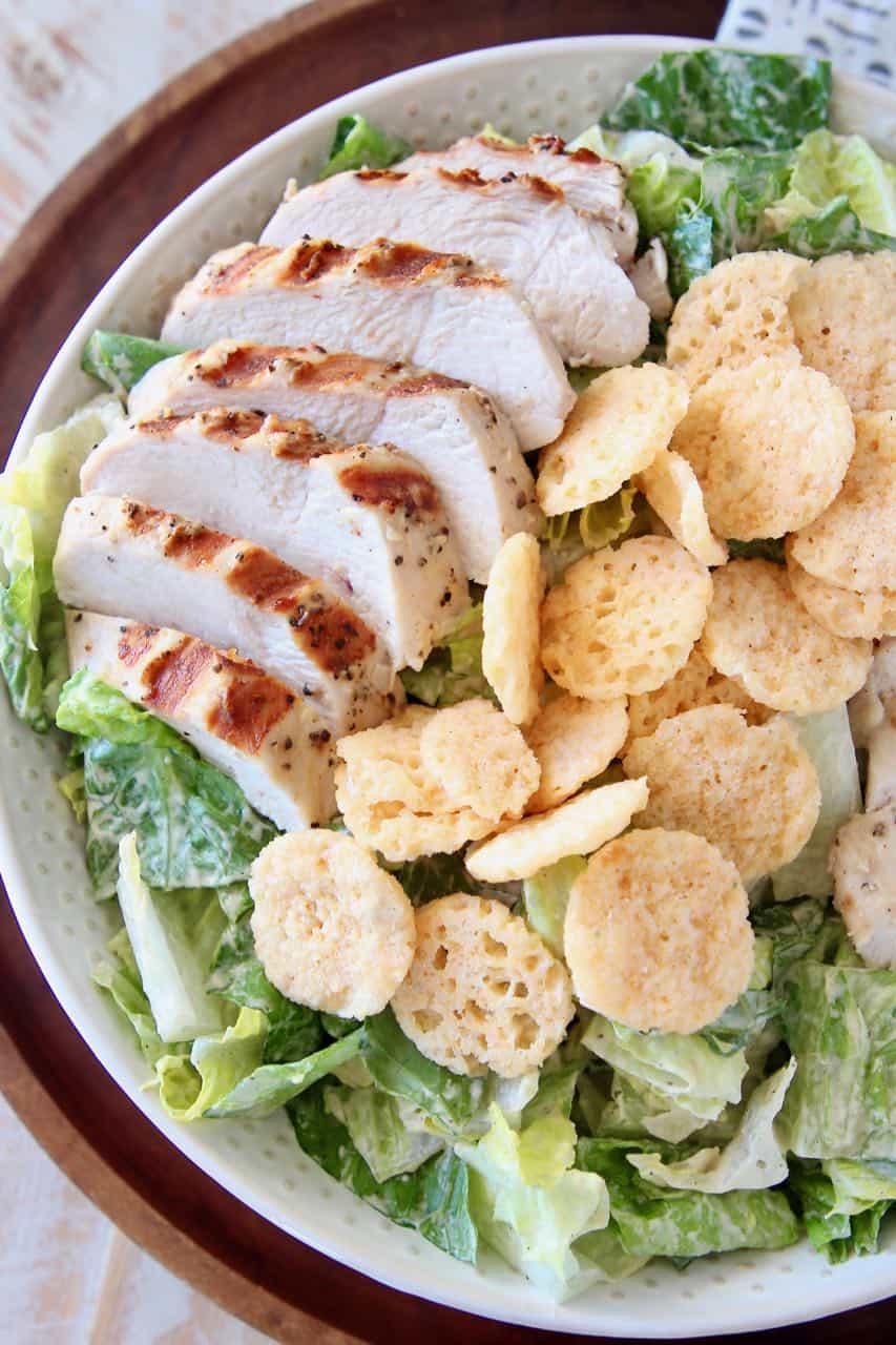 Bowl of caesar salad topped with sliced grilled chicken and parmesan cheese crisps