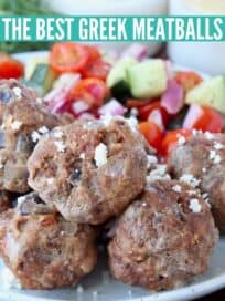 greek meatballs stacked up on plate next to tomato cucumber salad