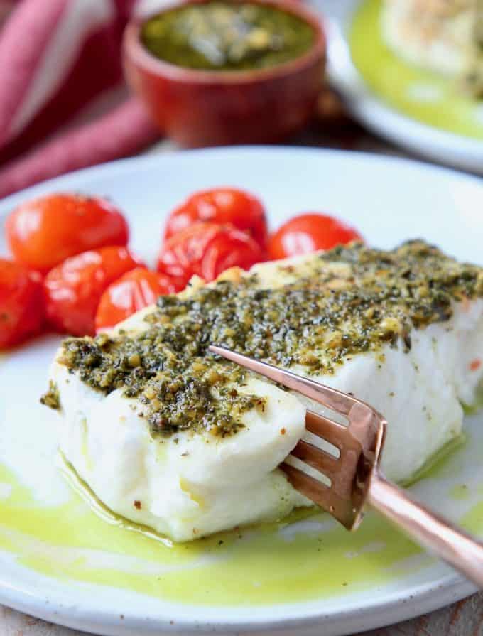 Fork cutting into sea bass on plate, topped with pesto