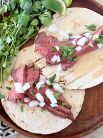 Two carne asada tacos on wood tray with fresh cilantro sprigs