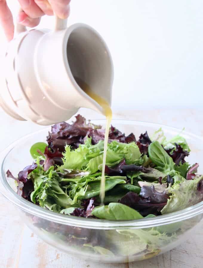 small pitcher pouring vinaigrette dressing on salad in a glass bowl