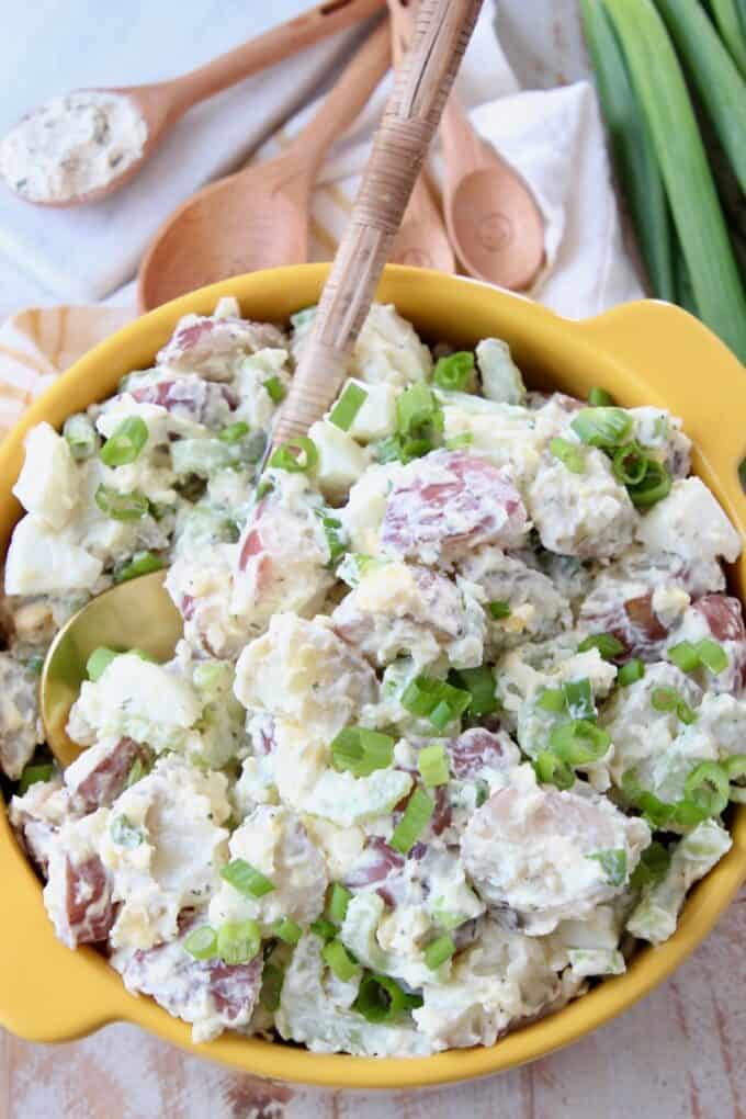 potato salad in yellow bowl with serving spoon and green onions on the side