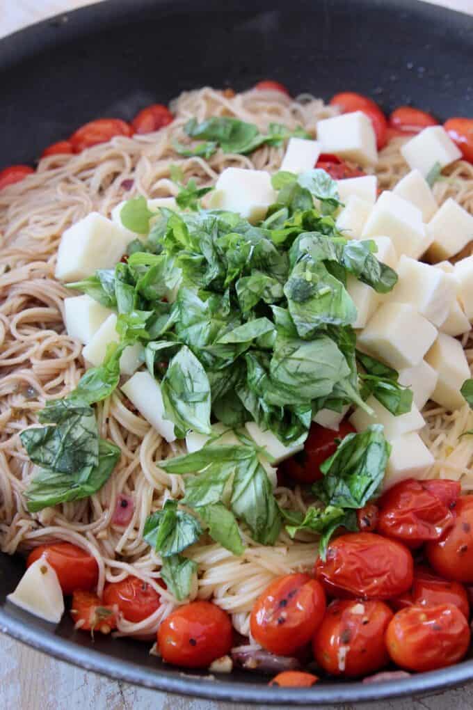 cooked angel hair pasta in a skillet topped with fresh basil, cubes of mozzarella cheese and cherry tomatoes