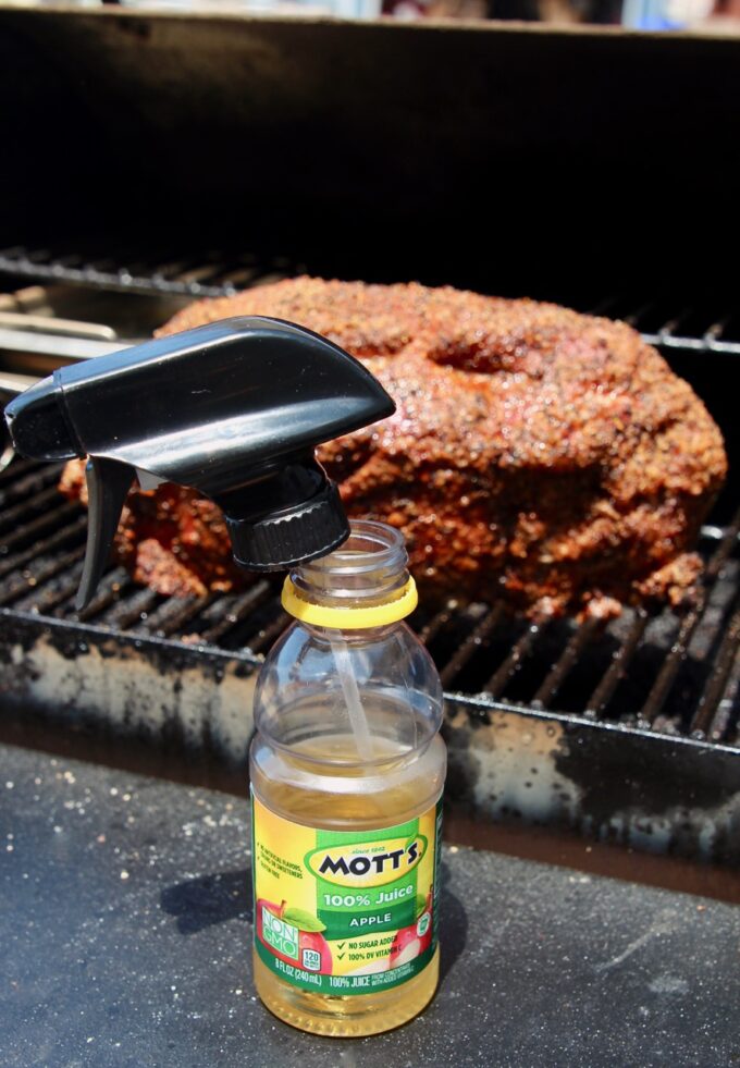 bottle of apple juice with a spray bottle lid in it in front of a pork shoulder on a smoker