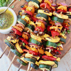 grilled sliced vegetables on skewers, stacked up on a wooden serving tray next to a bowl of sauce
