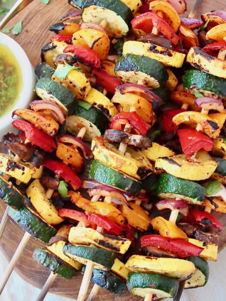 grilled sliced vegetables on skewers, stacked up on a wooden serving tray next to a bowl of sauce
