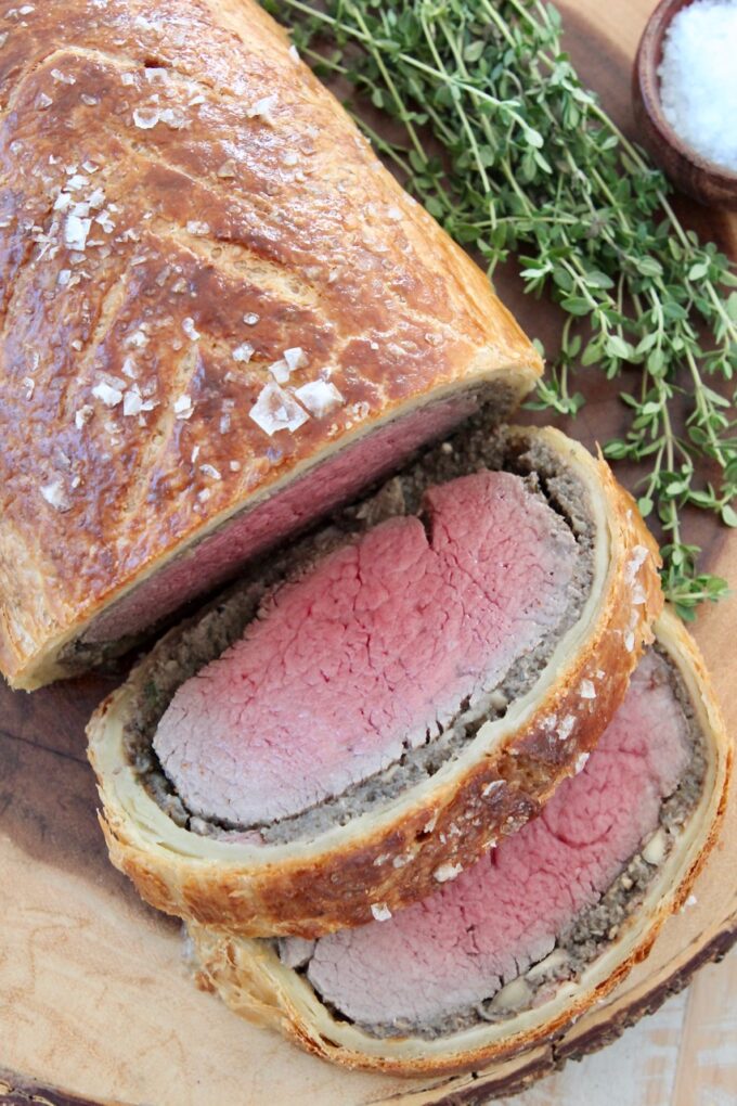 cooked sliced beef wellington on wood serving tray with fresh herbs