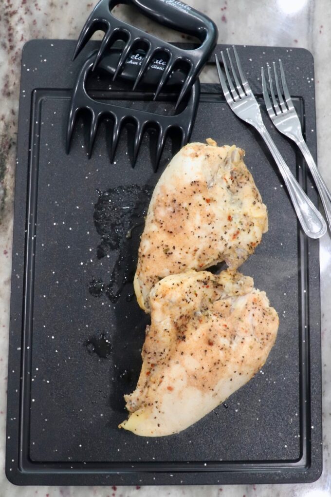 seasoned cooked chicken breasts on a cutting board with forks and meat claws on the side