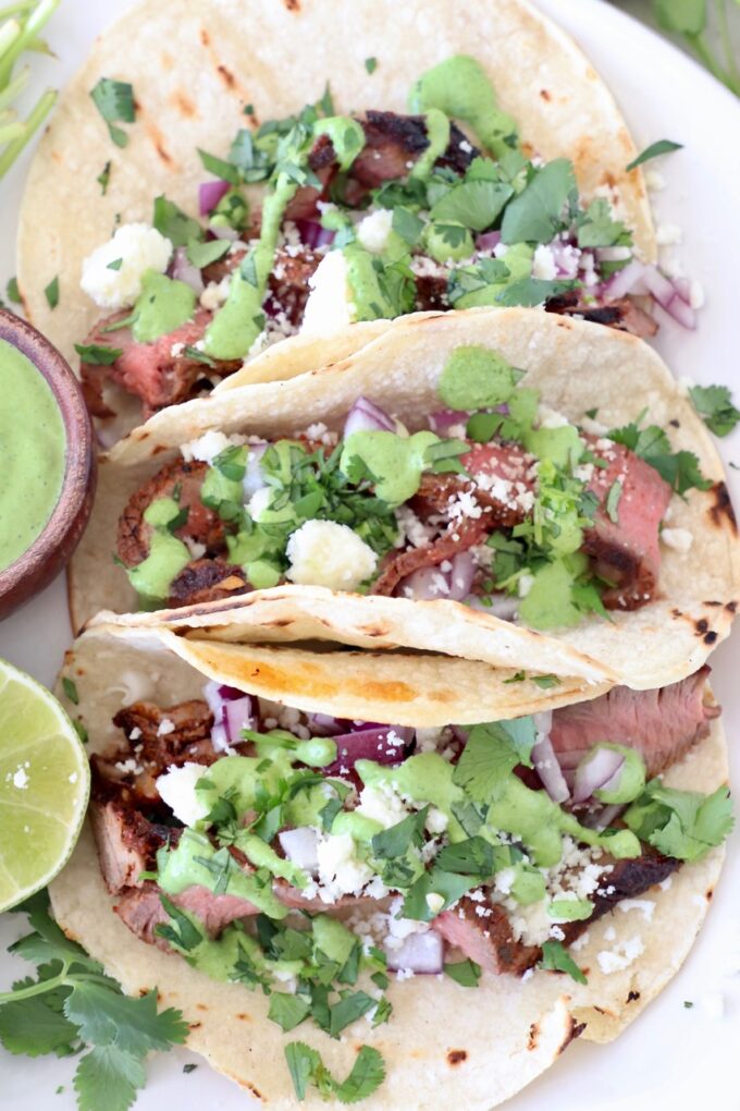 three steak tacos on plate topped with cilantro, sauce and cheese