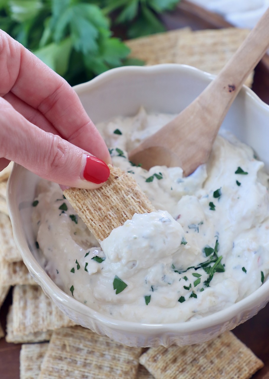 cracker dipped into garlic dip in bowl with wooden spoon