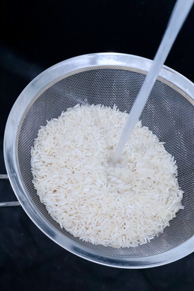 white rice in mesh strainer in sink with water pouring over the rice