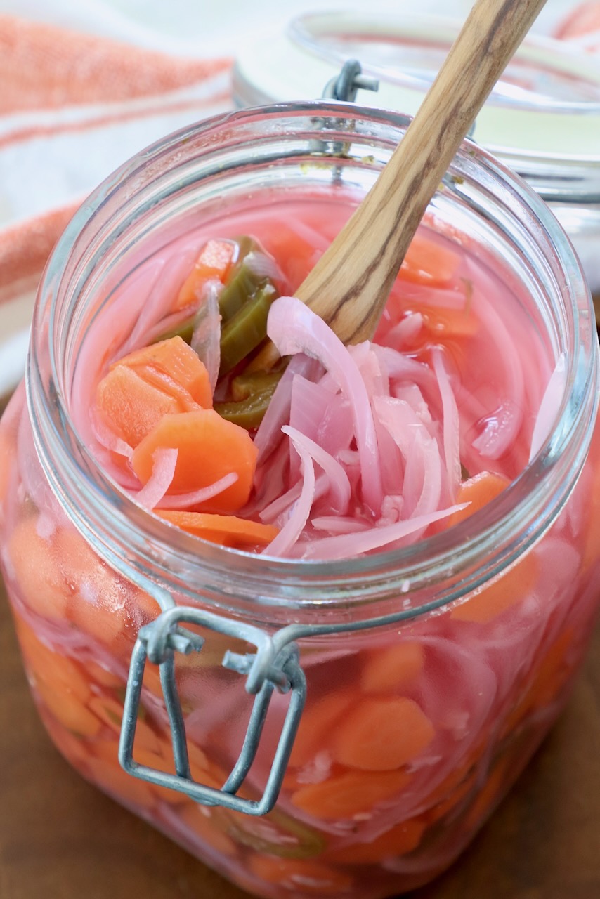 pickled sliced onions, carrots and jalapenos in large glass jar with wooden fork