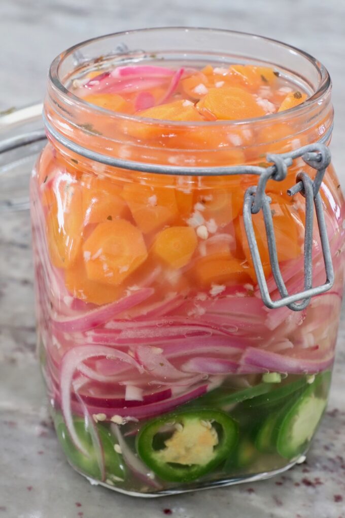 sliced carrots, onions and jalapenos in large glass jar covered in pickling liquid
