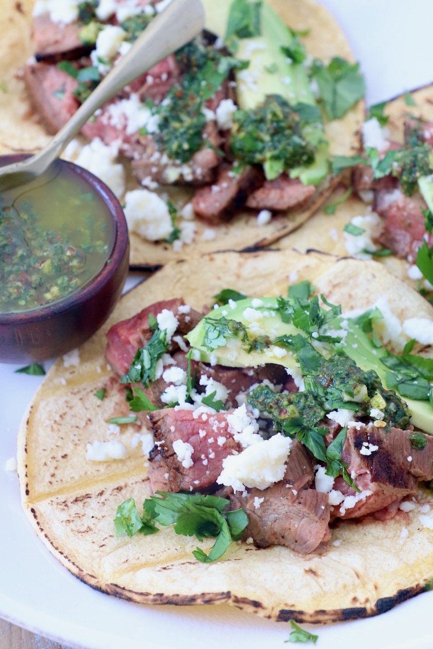 grilled steak in tortillas on plate topped with cotija cheese, avocado and chimichurri sauce