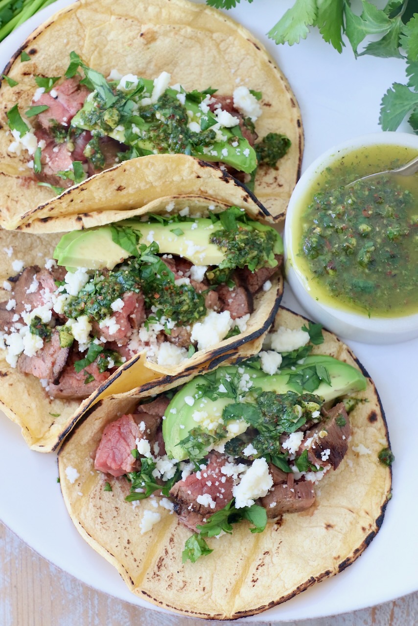 three tacos on plate filled with grilled steak, avocado and chimichurri sauce
