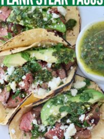 three tacos on white plate filled with grilled steak, cotija cheese, avocado slices and cilantro chimichurri sauce