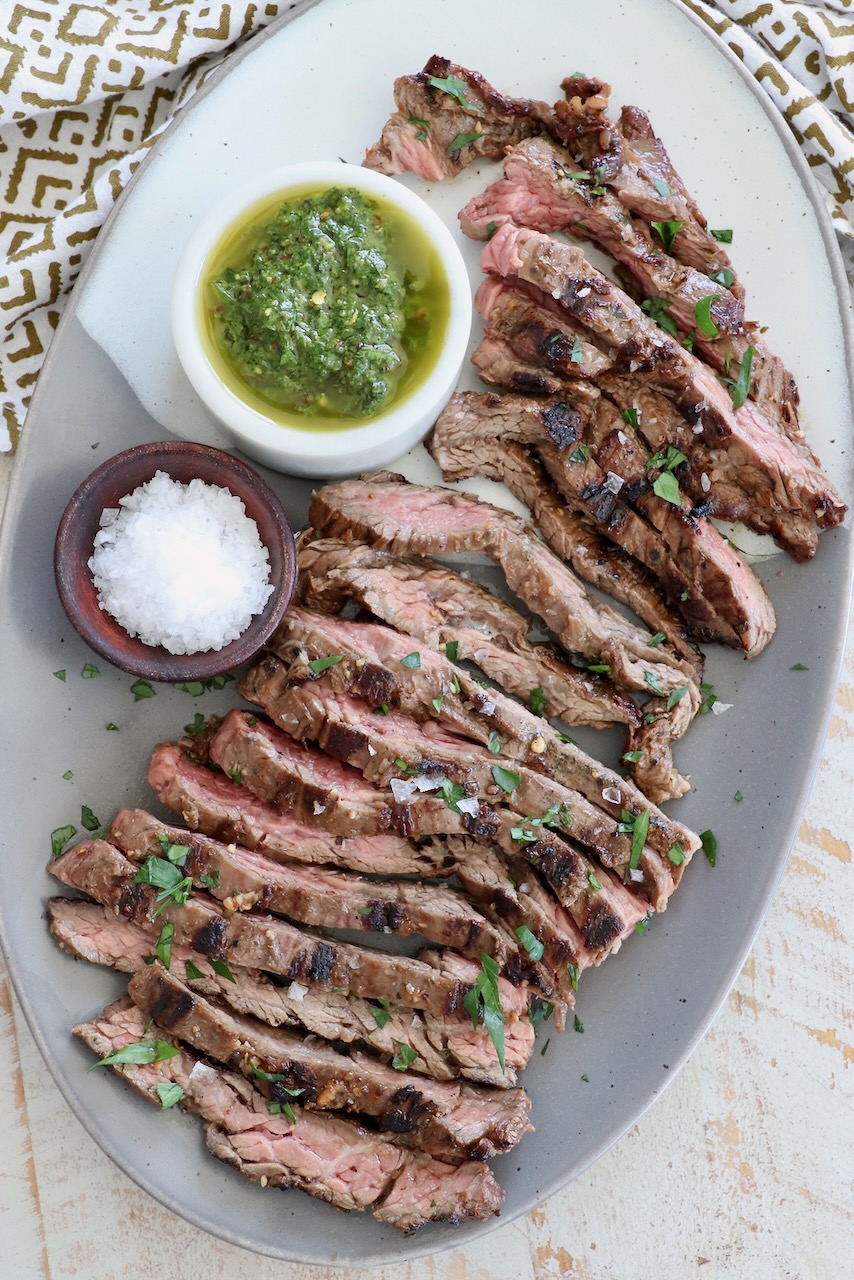 sliced grilled skirt steak on plate with small bowls of chimichurri sauce and flaky sea salt